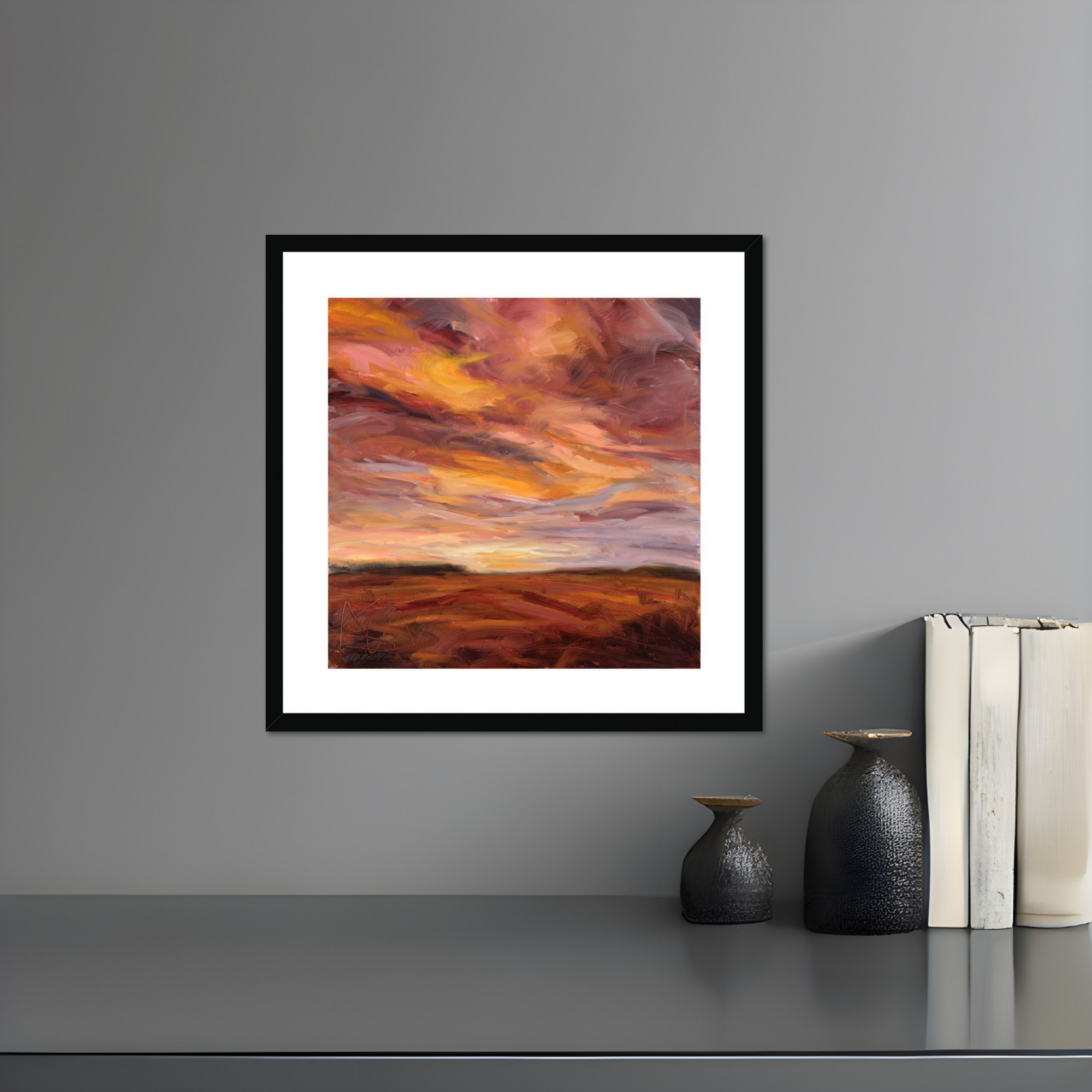 Framed abstract painting of a sunset over a field on a gray wall with decorative vases and books below.