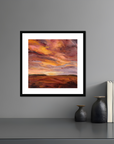 Framed abstract painting of a sunset over a field on a gray wall with decorative vases and books below.