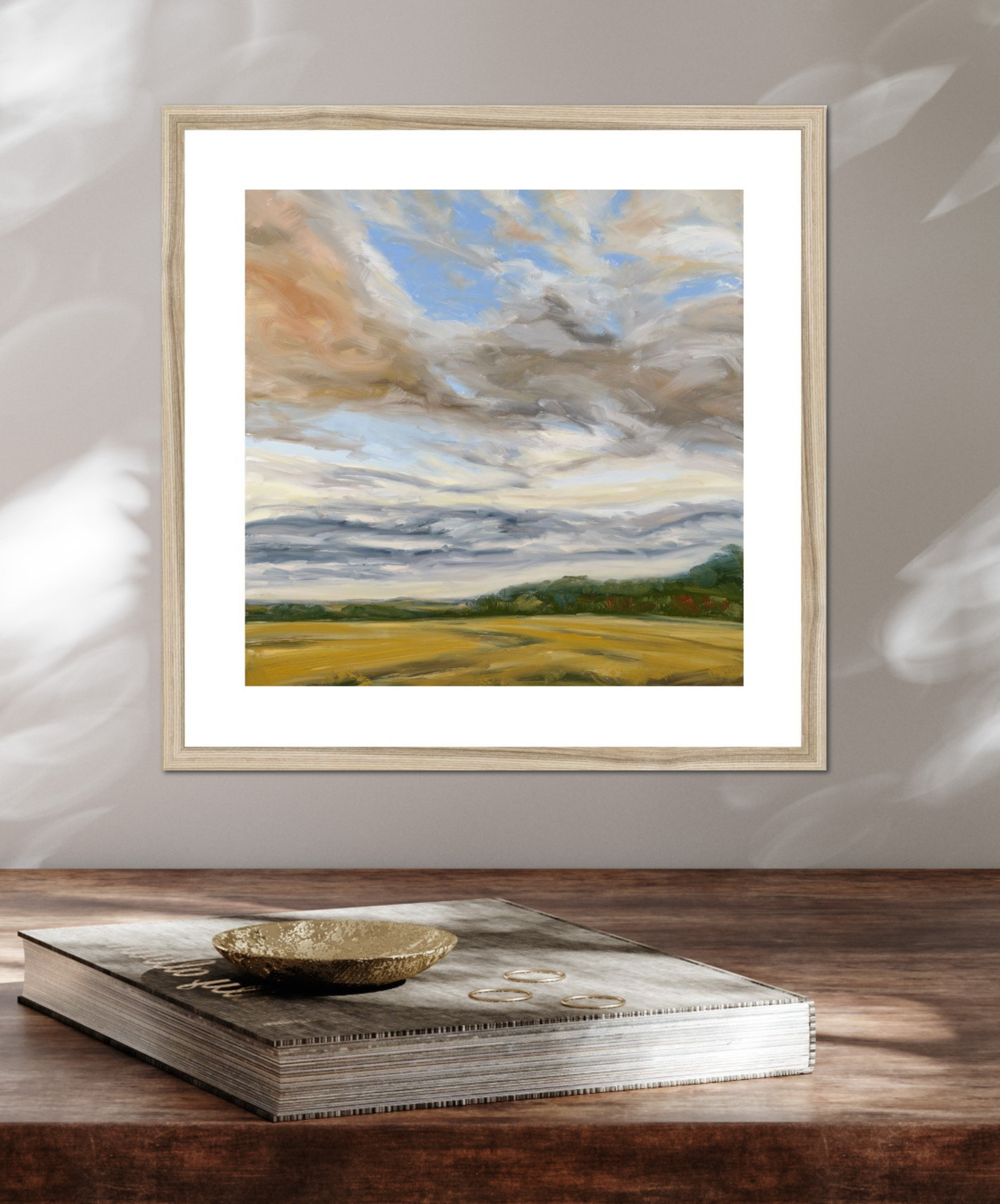 Framed painting of a landscape with clouds and fields on a wooden surface.