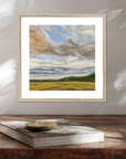 Framed painting of a landscape with clouds and fields on a wooden surface.