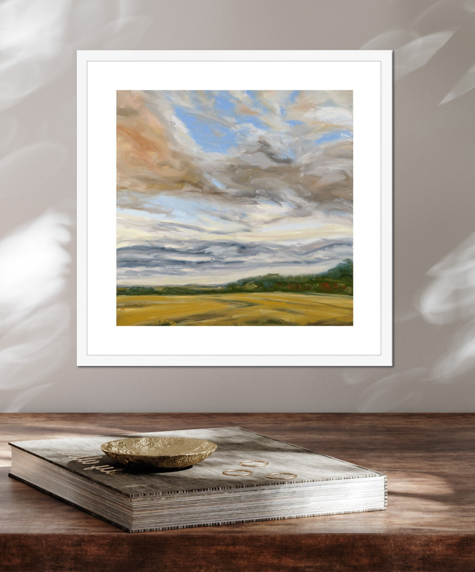 Framed painting of a landscape with clouds and fields on a wooden surface.