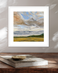 Framed painting of a landscape with clouds and fields on a wooden surface.
