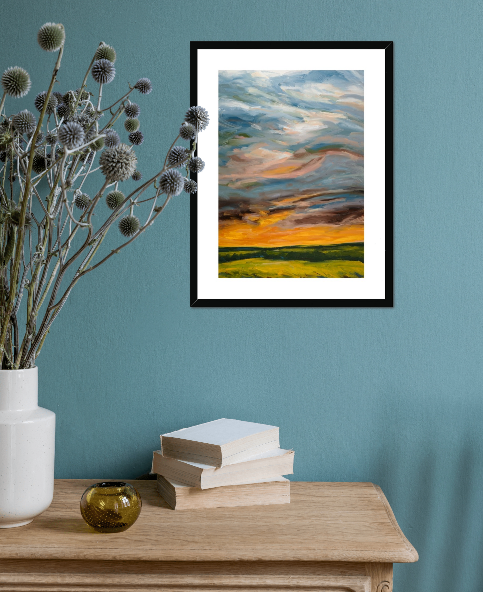 Framed artwork of a sunset over a field on a teal wall with a vase and books on a wooden surface.
