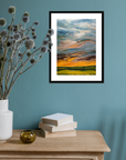 Framed artwork of a sunset over a field on a teal wall with a vase and books on a wooden surface.