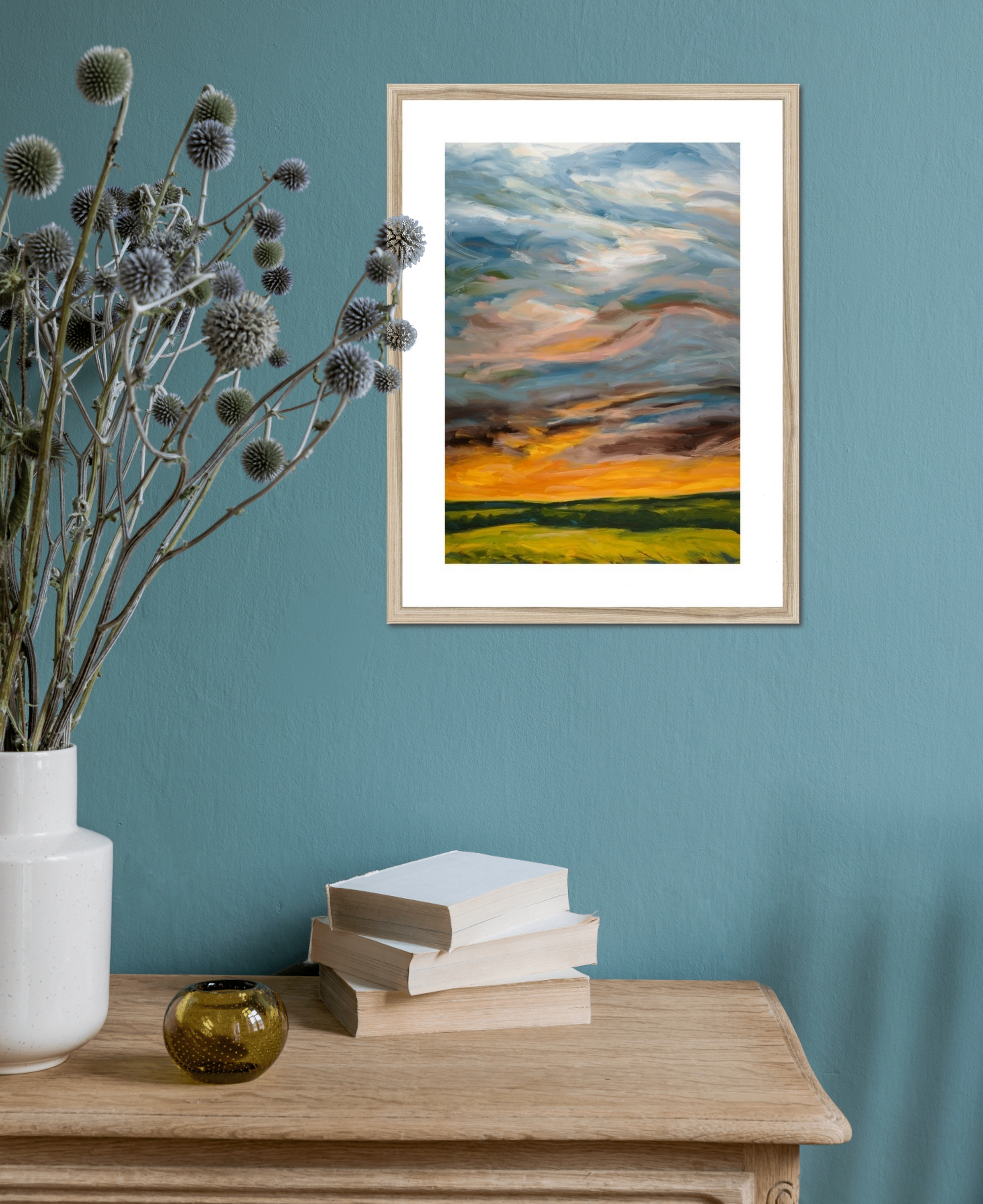 Framed artwork of a sunset over a field on a teal wall with decorative items on a wooden surface.