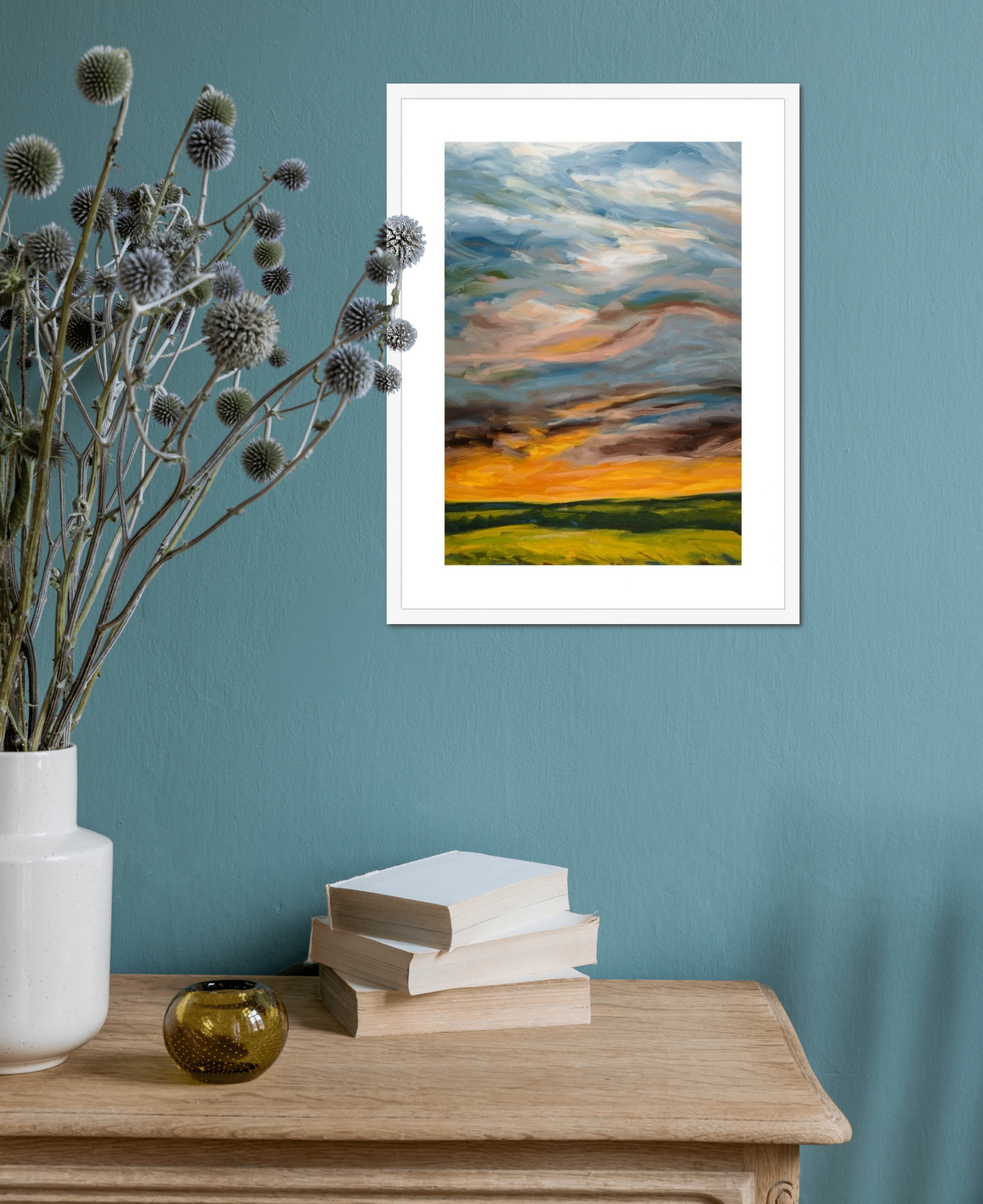 Framed artwork of a sunset over a field on a teal wall with decorative items on a wooden surface.