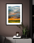 Framed abstract landscape painting on a wall with a candle and plant below.