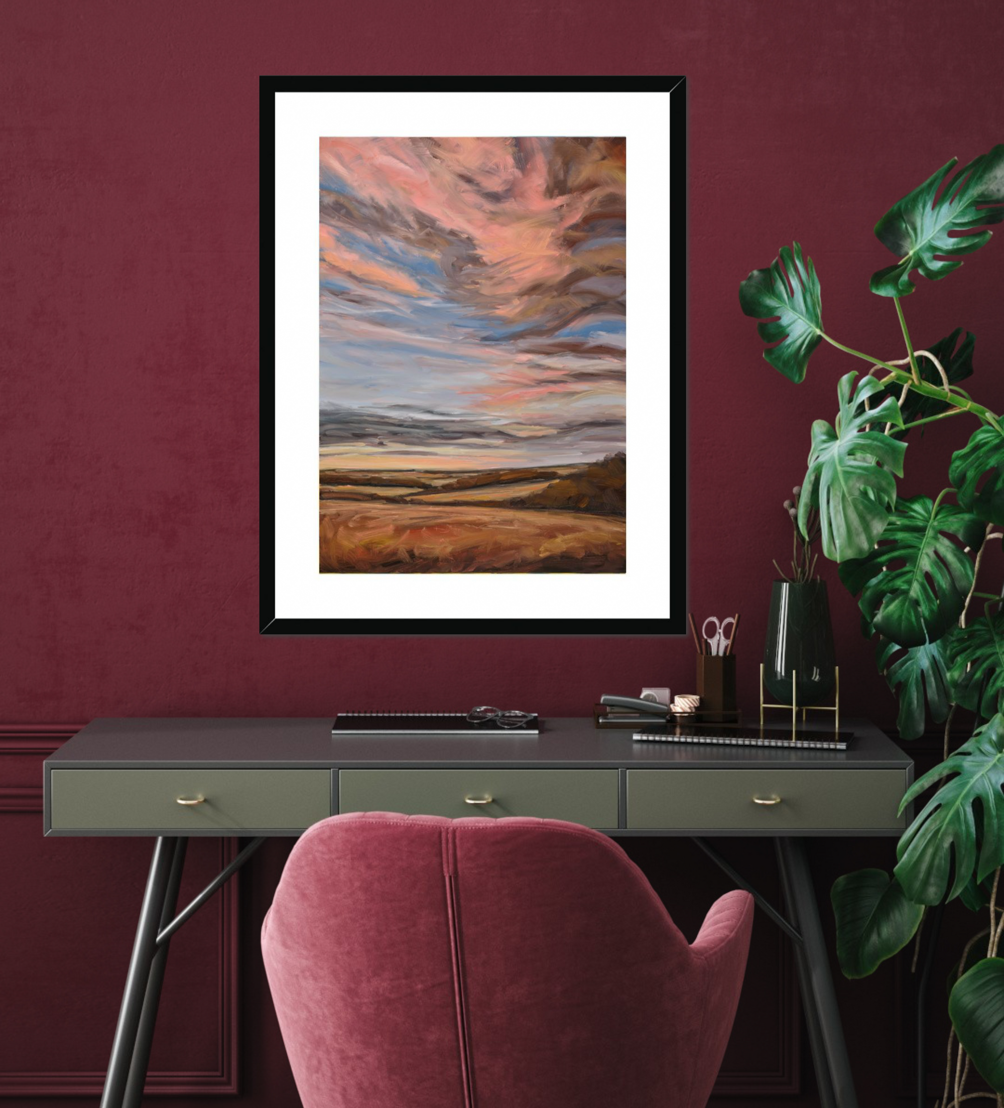 Framed abstract landscape art on a maroon wall above a desk with a pink chair.