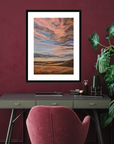 Framed abstract landscape art on a maroon wall above a desk with a pink chair.
