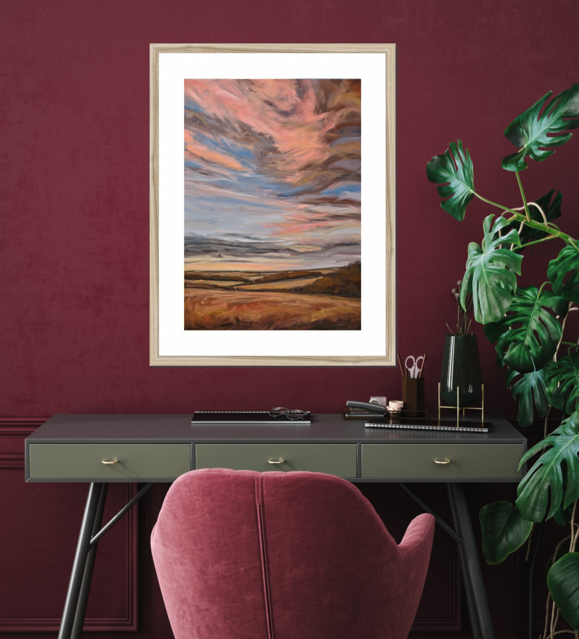 Framed abstract artwork on a wall above a desk with a pink chair and plant.