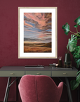 Framed abstract artwork on a wall above a desk with a pink chair and plant.
