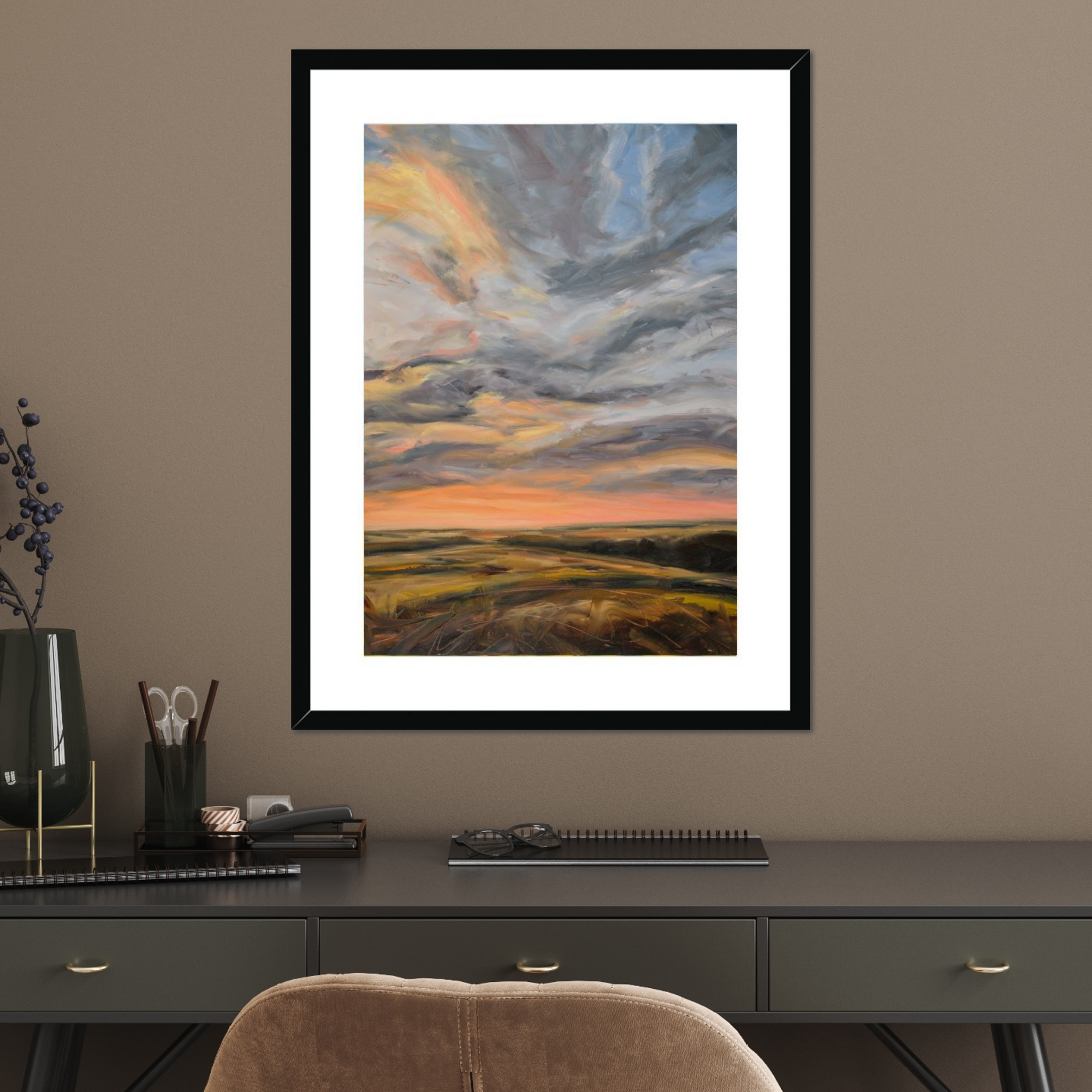 Framed artwork of a sunset over a field on a wall above a desk.