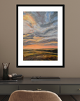 Framed artwork of a sunset over a field on a wall above a desk.