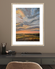 Framed artwork of a sunset over a field on a wall above a desk.