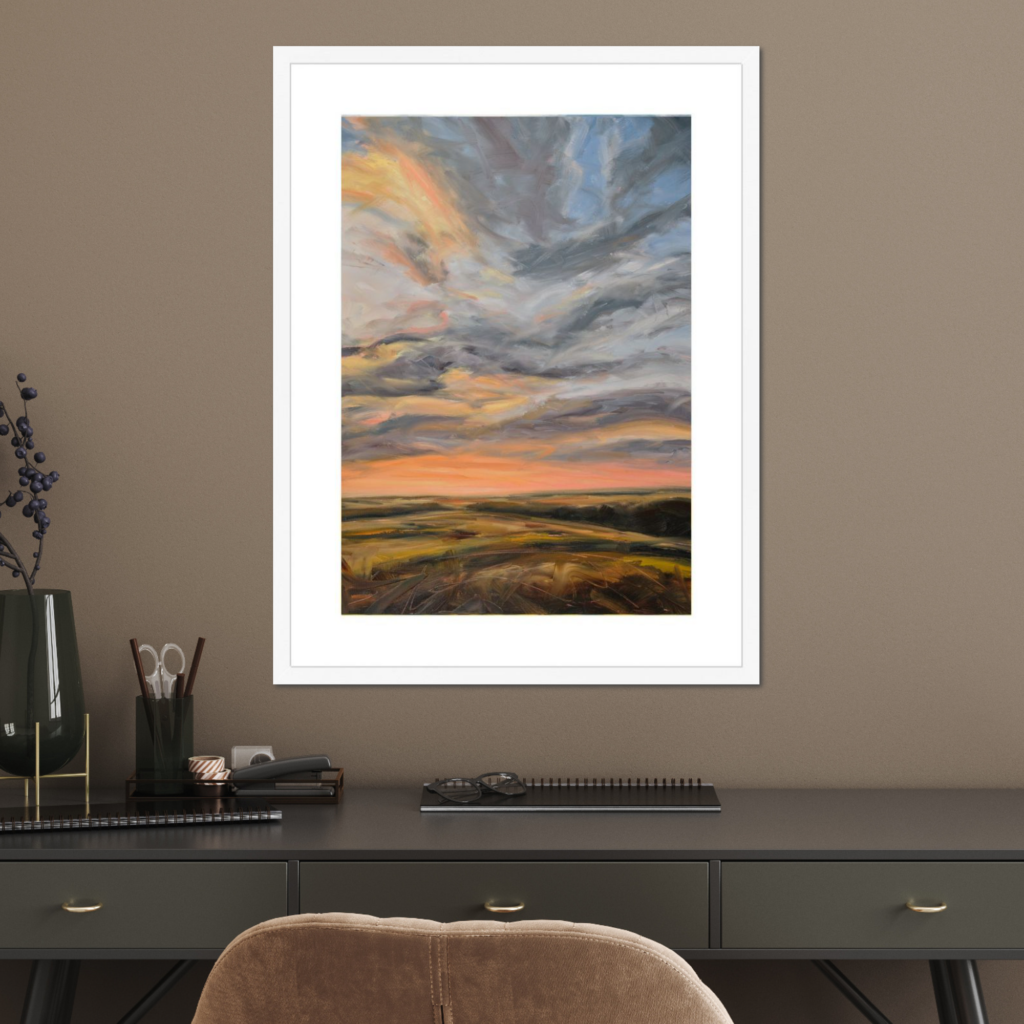 Framed artwork of a sunset over a field on a wall above a desk.