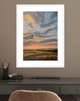 Framed artwork of a sunset over a field on a wall above a desk.