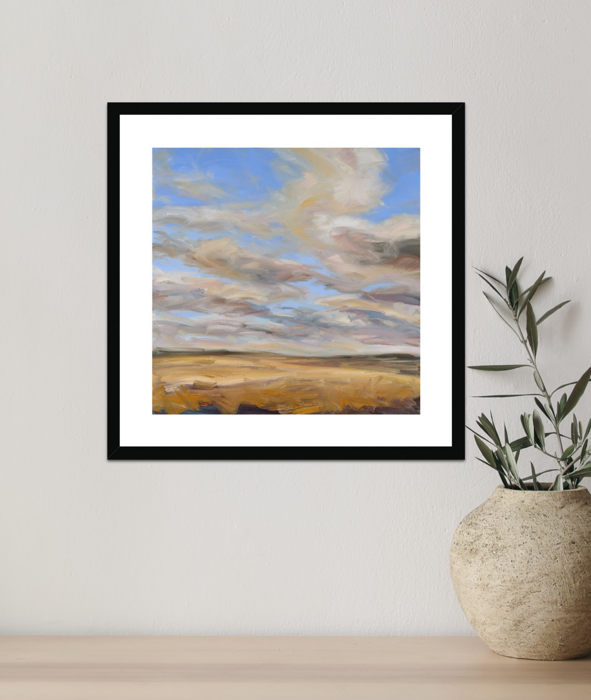 Framed artwork of a landscape with clouds on a wall next to a vase with plants.