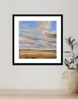 Framed artwork of a landscape with clouds on a wall next to a vase with plants.