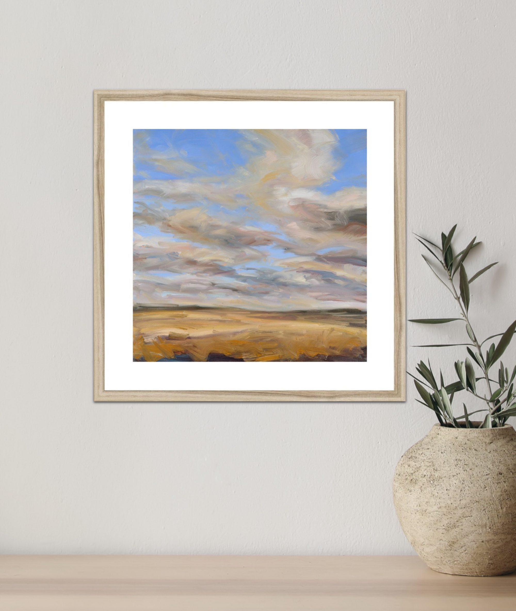 Framed artwork of a landscape with clouds on a wall next to a plant pot.