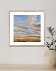Framed artwork of a landscape with clouds on a wall next to a plant pot.