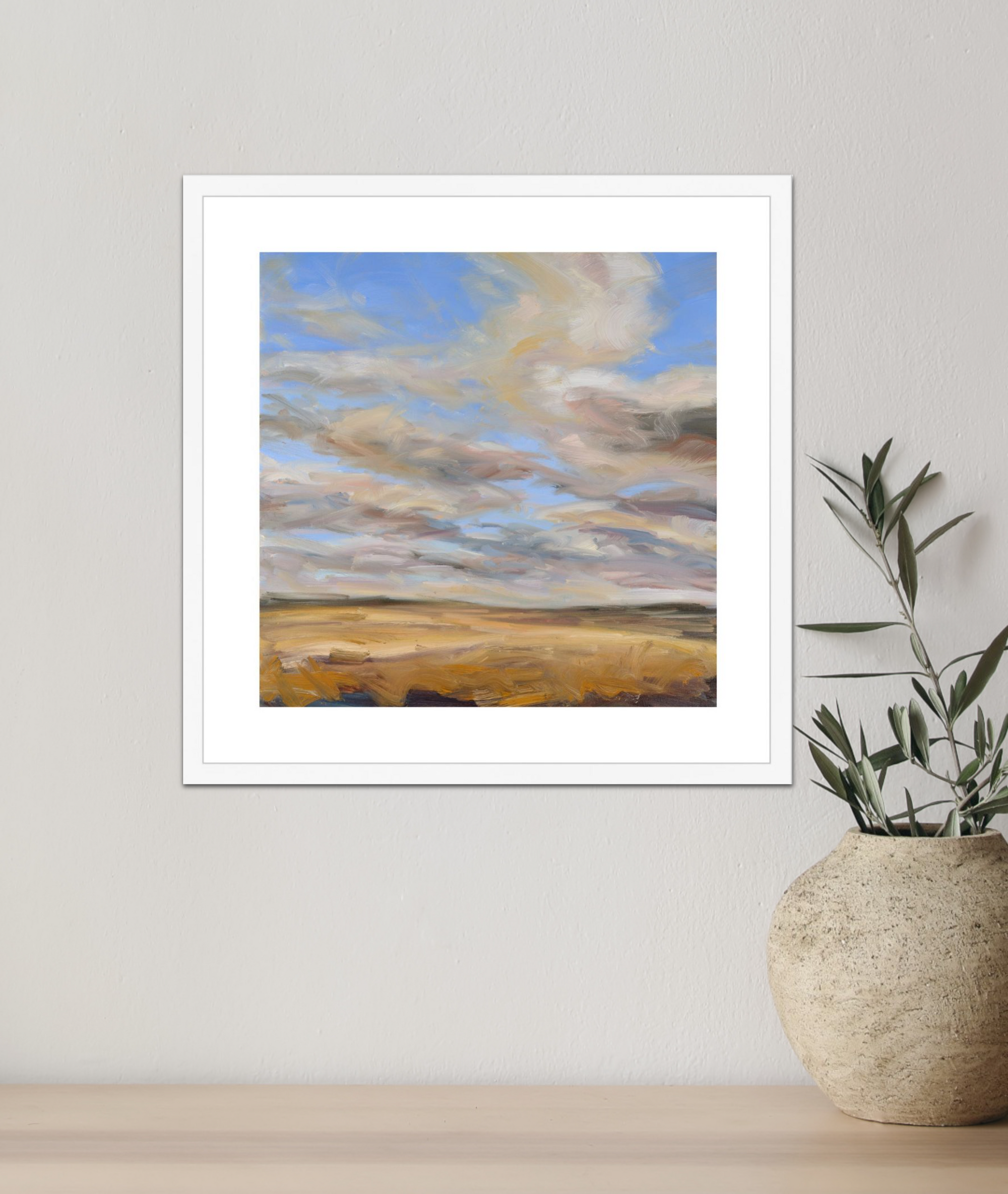 Framed artwork of a landscape with a blue sky and clouds, placed on a light wooden surface next to a textured vase.