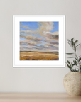 Framed artwork of a landscape with a blue sky and clouds, placed on a light wooden surface next to a textured vase.