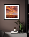 Framed artwork of a sunset over a landscape on a wall in a room with a cabinet and plant.