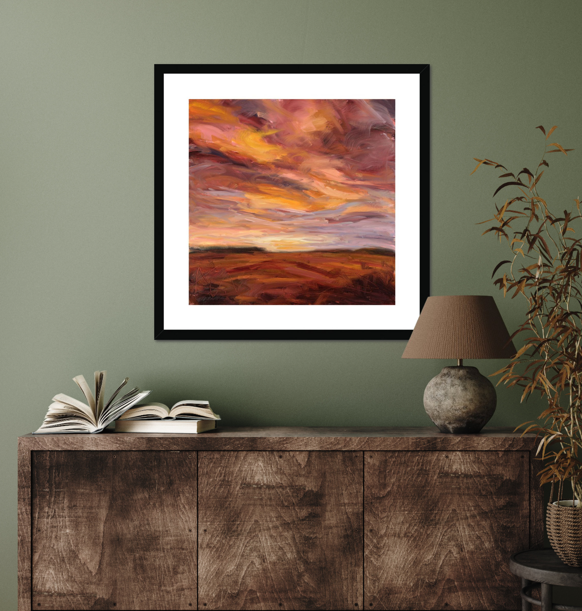 Framed artwork of a sunset over a landscape on a green wall above a wooden cabinet.