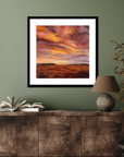 Framed artwork of a sunset over a landscape on a green wall above a wooden cabinet.