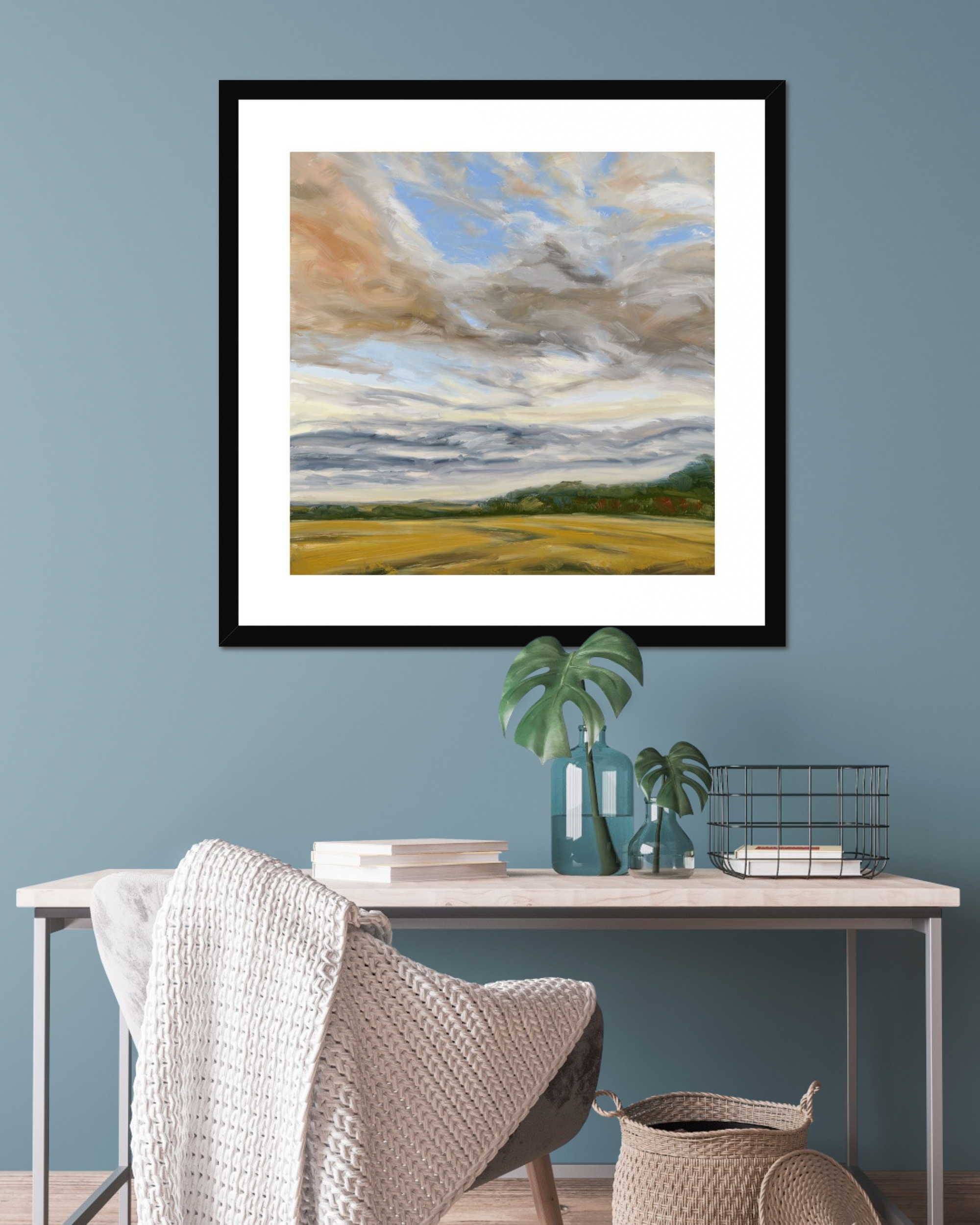 Framed landscape painting on a blue wall above a desk with decorative items.