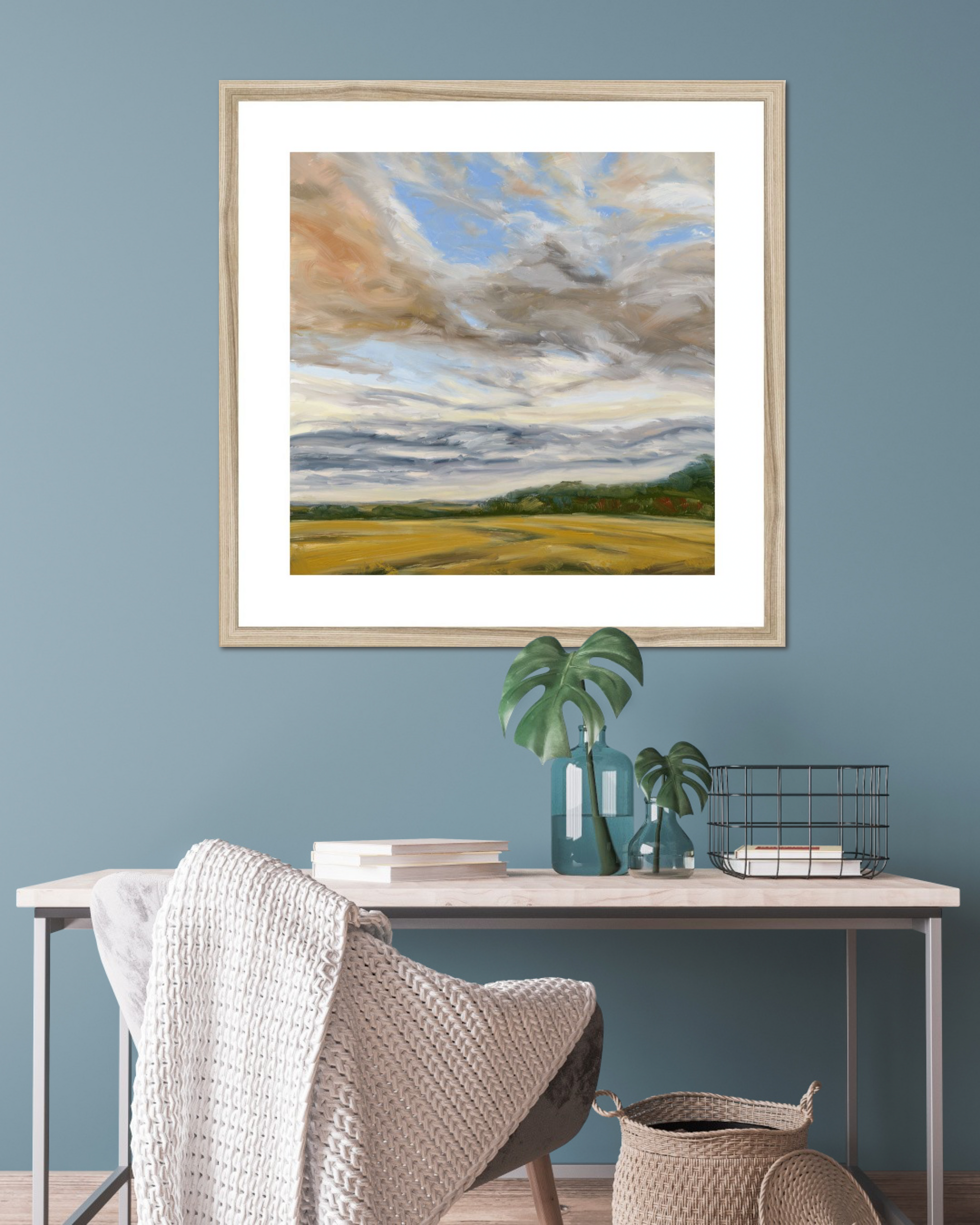 Framed landscape painting on a blue wall above a desk with decor items.