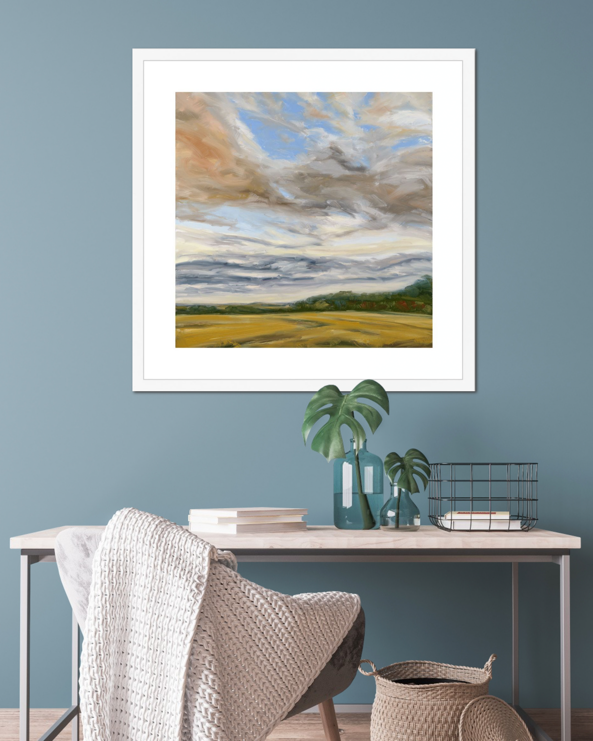 Framed landscape painting on a blue wall above a desk with decorative items.