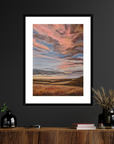 Framed artwork of a sunset over a field on a dark wall above a wooden console table.