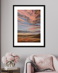 Framed abstract painting on a wall above a pink armchair with a small table and flowers.