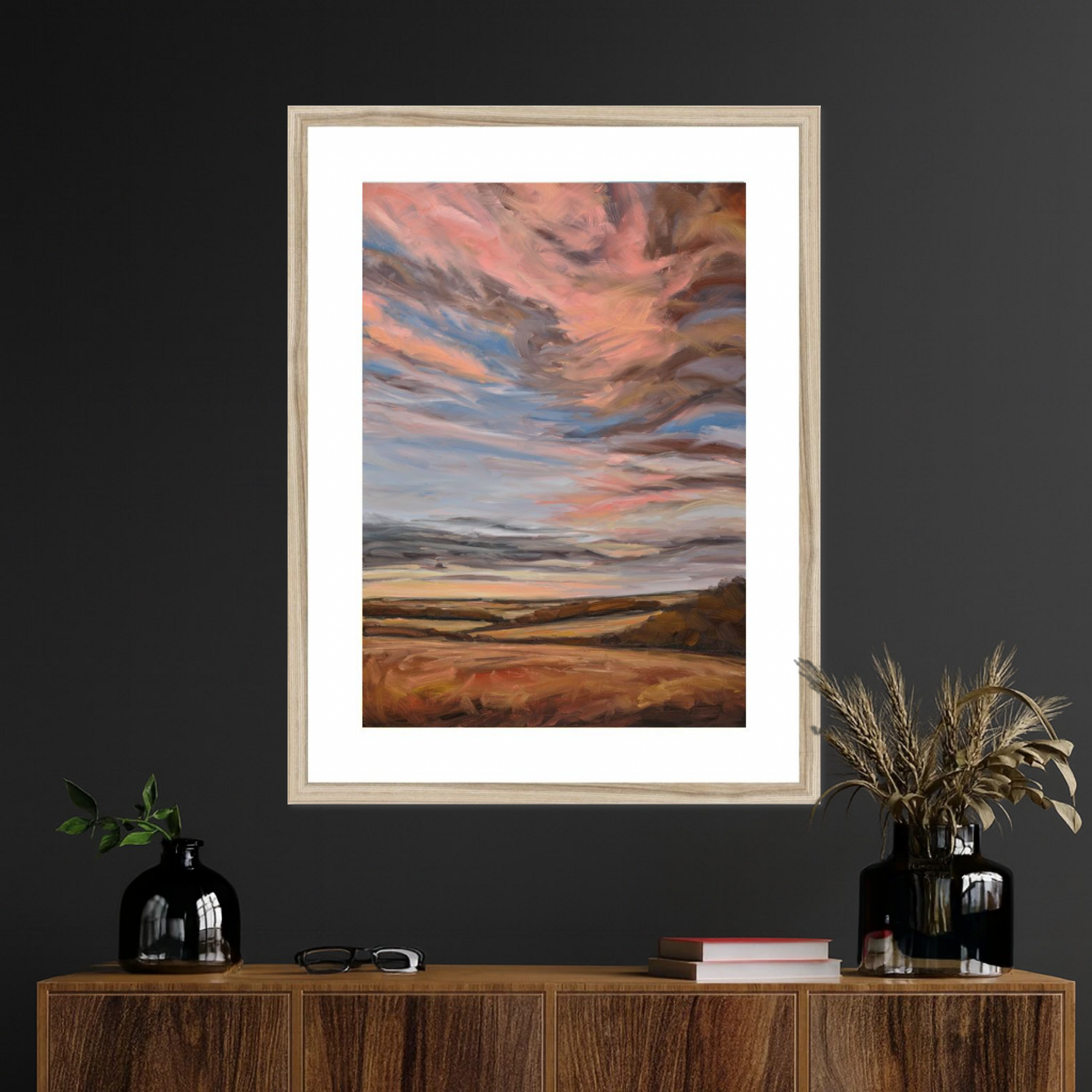 Framed artwork of a sunset over a landscape on a dark wall above a wooden console table.