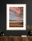 Framed artwork of a sunset over a landscape on a dark wall above a wooden console table.