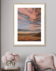 Framed artwork of a landscape with pink and orange hues on a gray wall above a pink armchair.