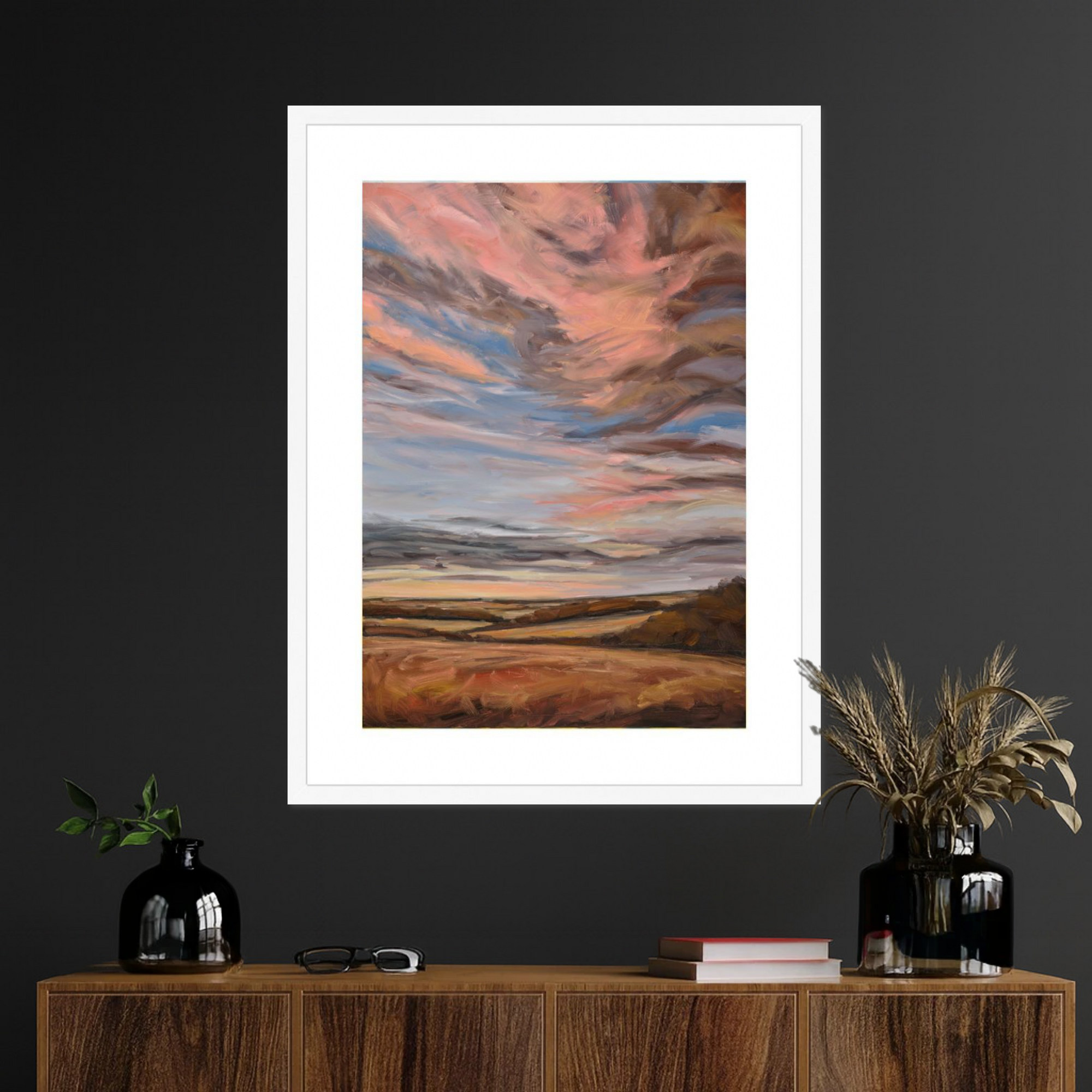 Framed artwork of a sunset over a landscape on a dark wall above a wooden console table.