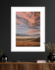 Framed artwork of a sunset over a landscape on a dark wall above a wooden console table.