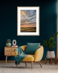 Yellow armchair with teal cushions and a green blanket in a room with a framed landscape painting on the wall.