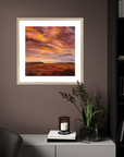 Framed artwork of a sunset over a landscape on a wall with decorative elements below.